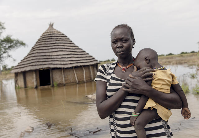 Abuk holds her child, Nyirou, outside of their flooded home. Water has flooded the area surrounding the home.